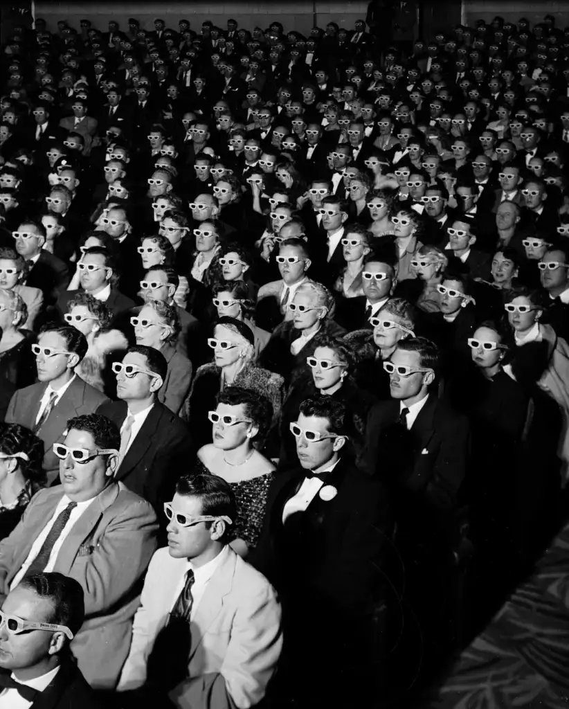 A large audience sits in a theater, all wearing white 3D glasses, watching an unseen screen. The crowd spans from the foreground to the background, filling the entire space. The image is in black and white, suggesting a historical context.