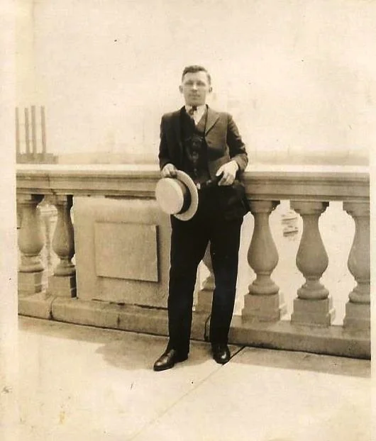 A man in a vintage suit leans against a stone balustrade, holding a straw boater hat. The photo has a sepia tone, giving it an old-fashioned look. The background is bright, suggesting a daytime setting.