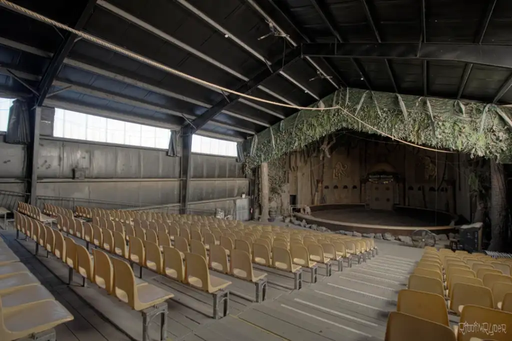 Interior view of an empty, rustic theater with rows of yellow seats facing a small wooden stage. The stage is surrounded by faux greenery, and the canopy is dark with exposed beams.