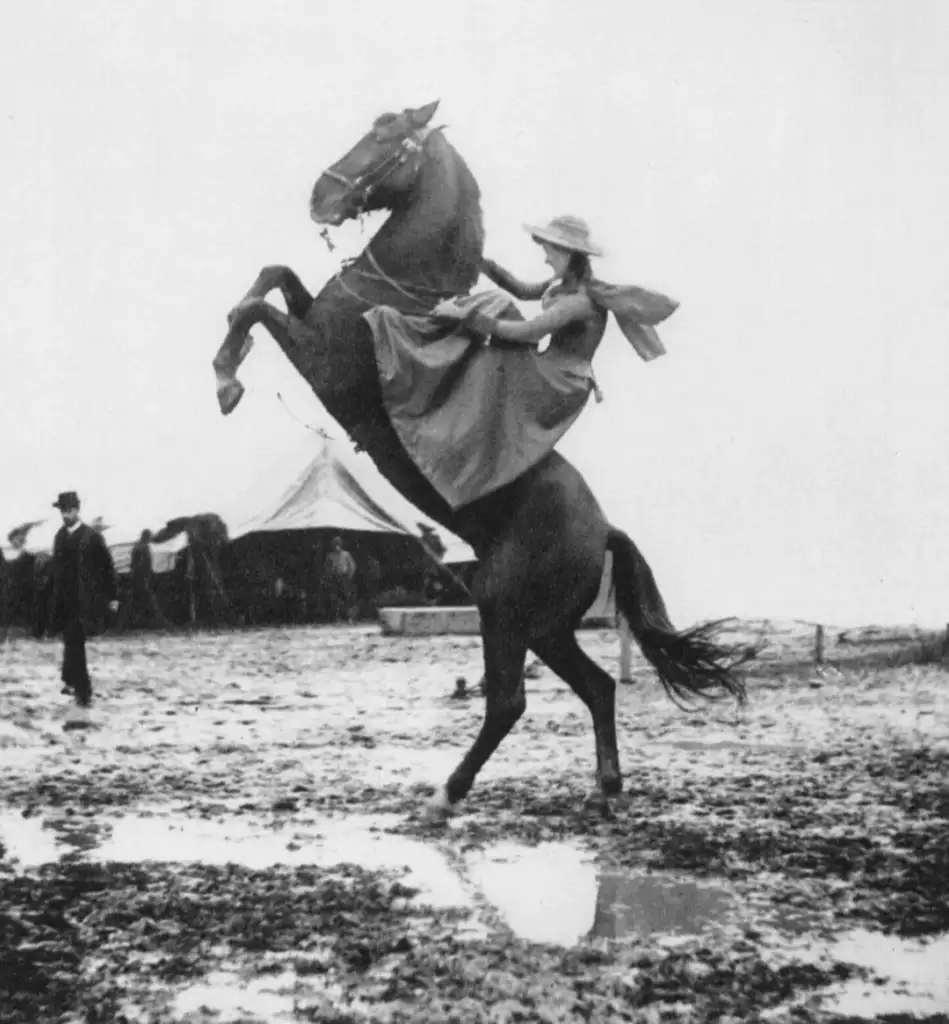 A woman wearing a dress and hat rides a rearing horse in a muddy field. In the background, a tent and several people are visible, with one person standing and observing the scene. The sky is overcast, adding to the dramatic effect.