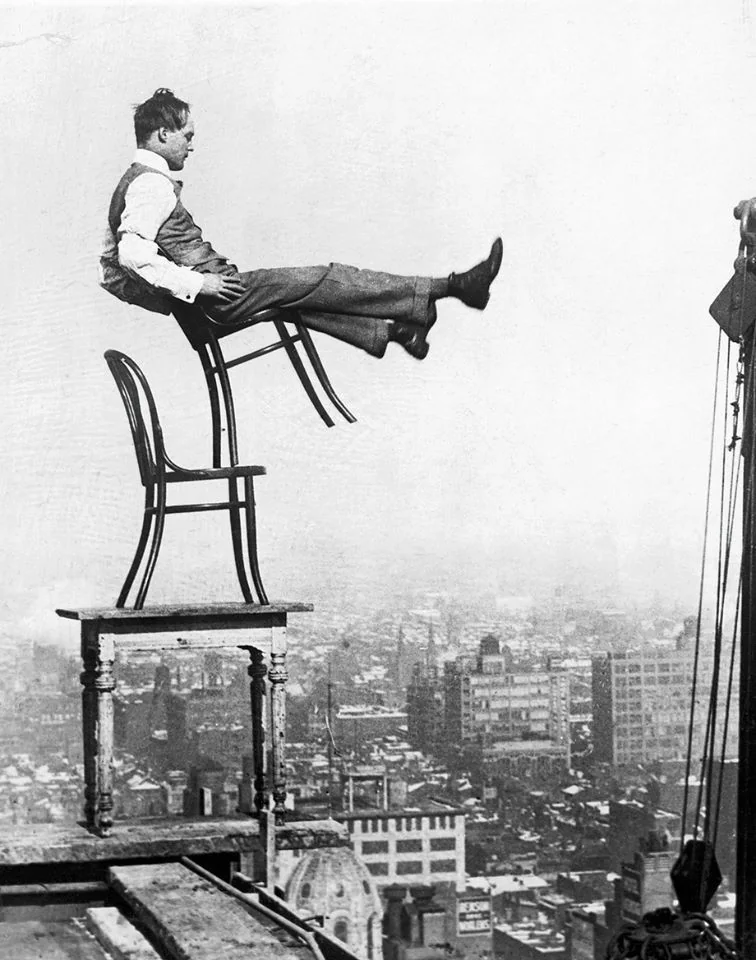 A man in a suit and tie balances on two chairs and a table atop a rooftop, his legs extended in the air. The background shows a hazy skyline with various buildings. The situation appears precarious and daring.