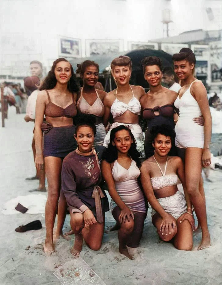 A group of eight women posing on a beach in vintage swimwear. Some are kneeling and others standing, all smiling. The background shows a sandy beach and people in the distance. The image has a retro, colorful aesthetic.