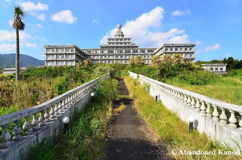 A path leads to a large, abandoned building surrounded by overgrown vegetation. The building has multiple stories and a central dome. White balustrades line the path. A single palm tree stands to the left under a sunny sky with clouds.