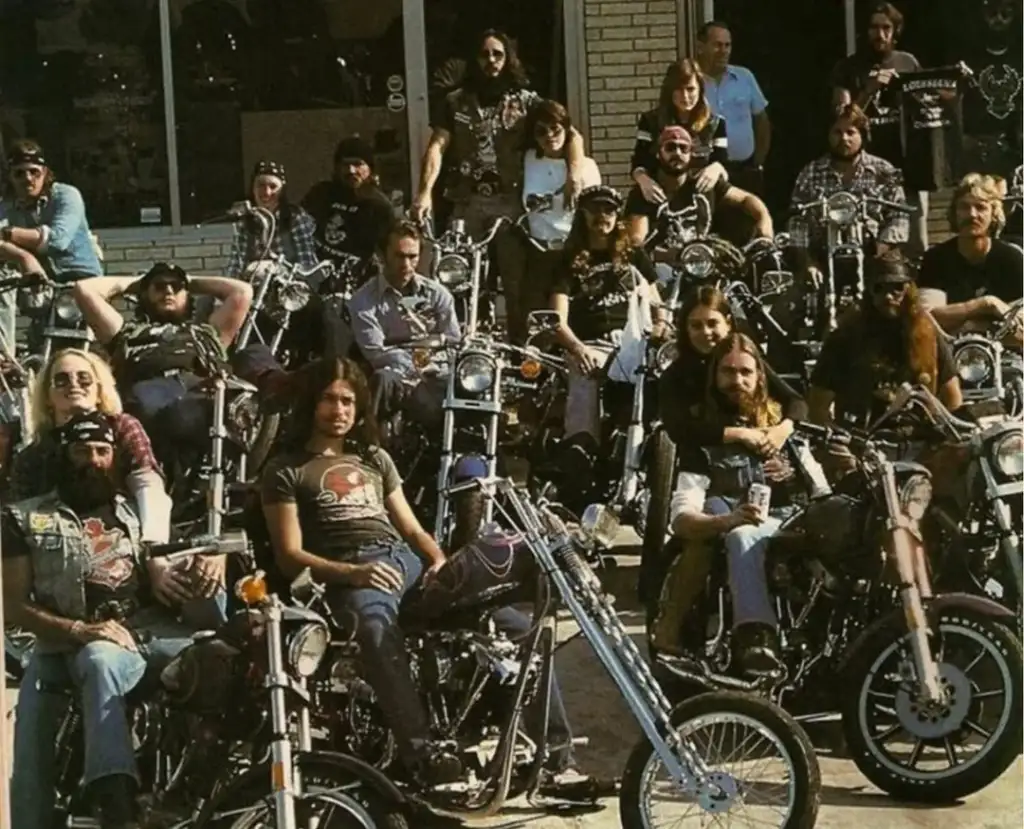 A group of people sit on motorcycles gathered in front of a building. The scene includes individuals with long hair and beards, wearing casual clothing like denim and graphic tees, evoking a biker culture atmosphere.