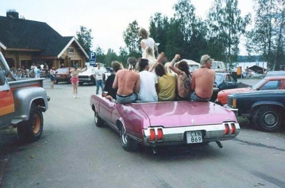 A group of people are sitting on the back of a pink convertible car parked in a lot. The scene is set in a rural area with trees in the background, and several other vehicles and people are visible around them.