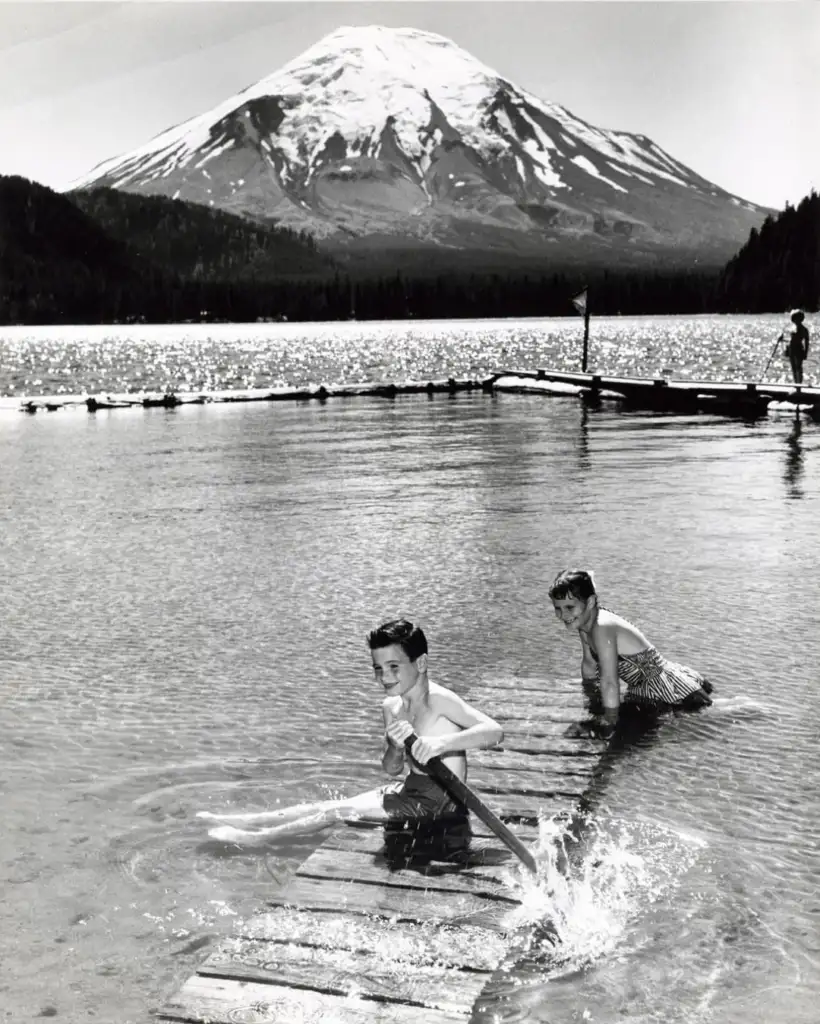 Two children play on a wooden dock in a lake, splashing water. In the background, a snow-capped mountain rises under a clear sky. Other figures can be seen relaxing by the lake. The scene is serene and sunlit.