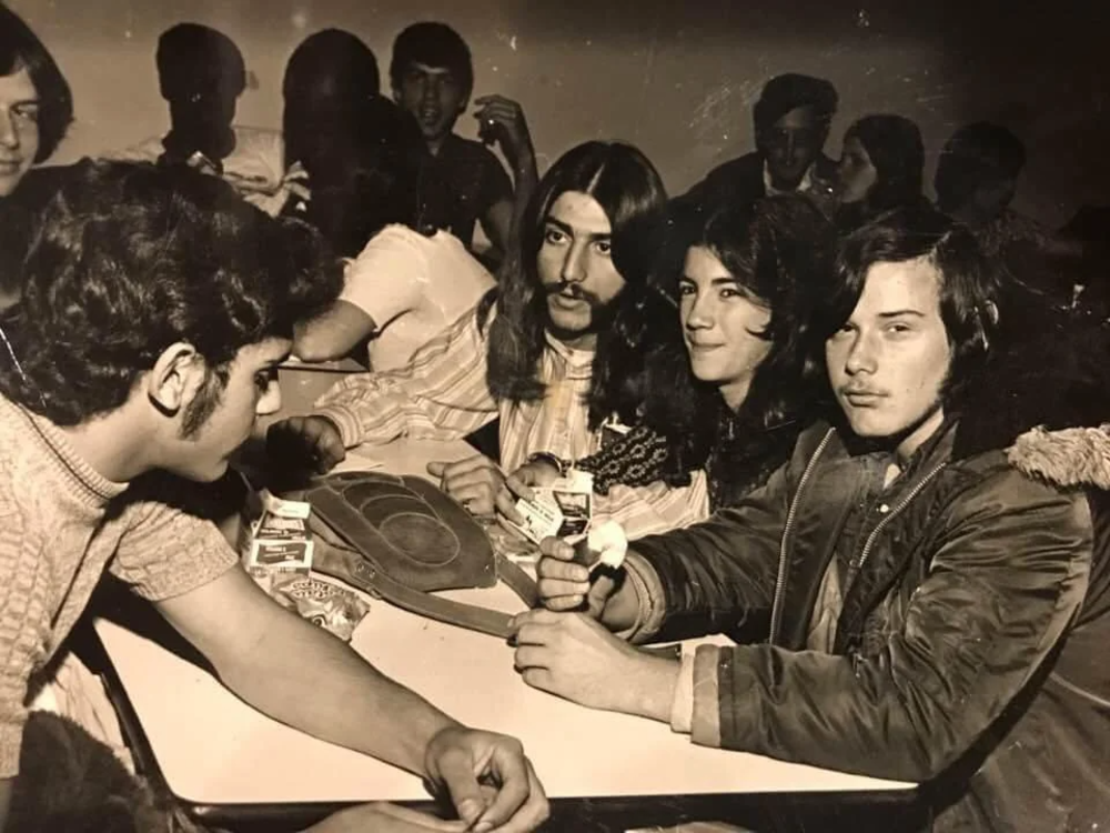 A black and white photo shows a group of young men seated around a table. They are engaging with each other, holding trading cards, and a circular board game is on the table. The background has more people sitting at other tables.