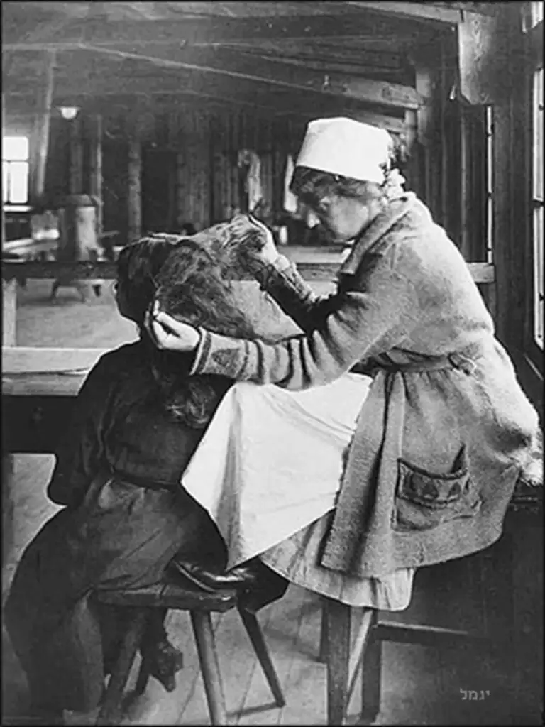 A person wearing a white headscarf and coat examines a child's head for lice indoors. The child is seated with their back to the camera. The background features large windows and wooden walls.