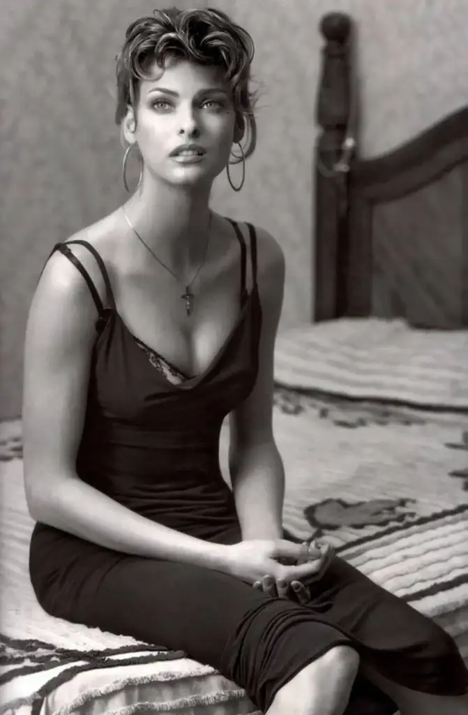 A woman in a sleeveless black dress sits on a bed with a patterned blanket. She has short, styled hair and wears large hoop earrings and a necklace with a cross pendant. The background features a wooden bedpost and a textured wall.