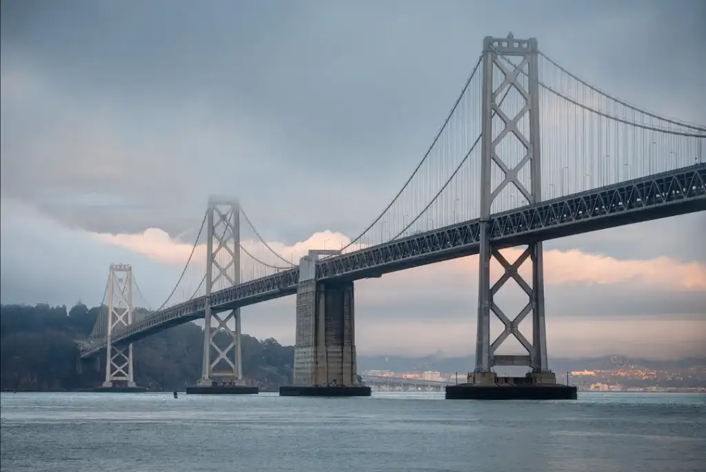 A large suspension bridge with multiple towers extends over a calm body of water. The bridge is partially shrouded in fog, with hills in the background and a cityscape visible beneath a cloudy sky.