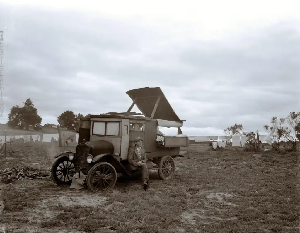 A man sits on the running board of an old truck with a makeshift wooden camper shell. The vehicle is parked in a field with clothes hanging on a line in the background under a cloudy sky. Sparse trees are visible in the distance.