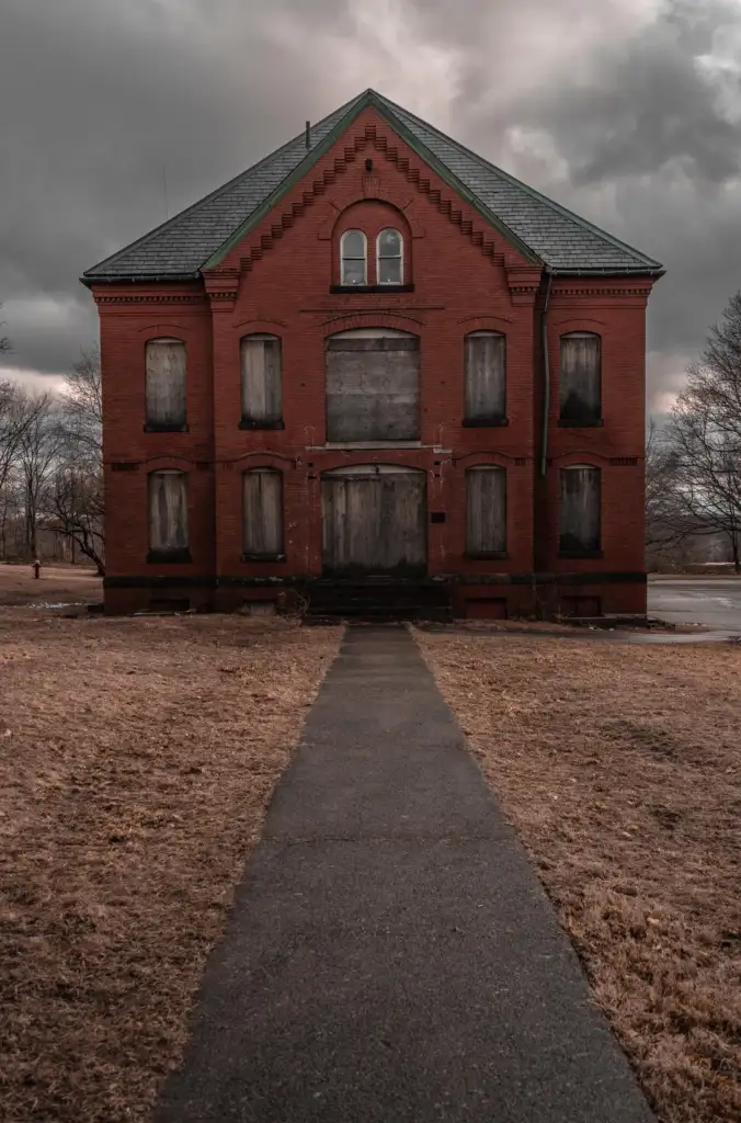 A gloomy, abandoned brick building with boarded-up windows and a closed wooden door. The structure is set against an overcast sky, with an empty pathway leading up to it through a barren field.