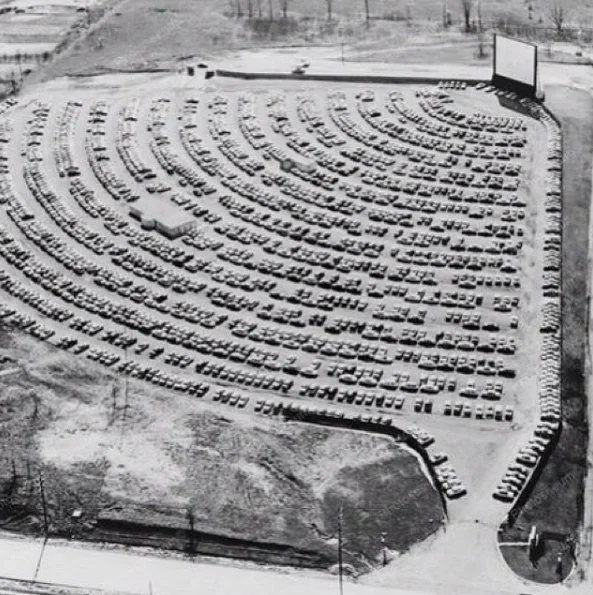 An aerial view of a vintage drive-in theater filled with numerous cars parked in semi-circular rows, all facing a large movie screen. The theater is surrounded by grassy and dirt areas.