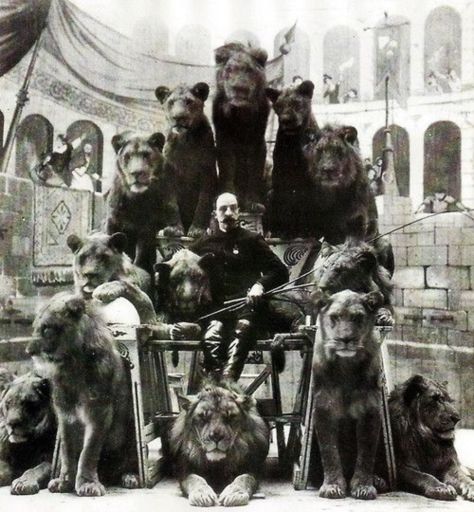 A man in a vintage circus setting sits confidently in the center of a group of ten lions. The lions surround him on various levels, with some seated on platforms. The backdrop shows a decorated interior with spectators in the distance.