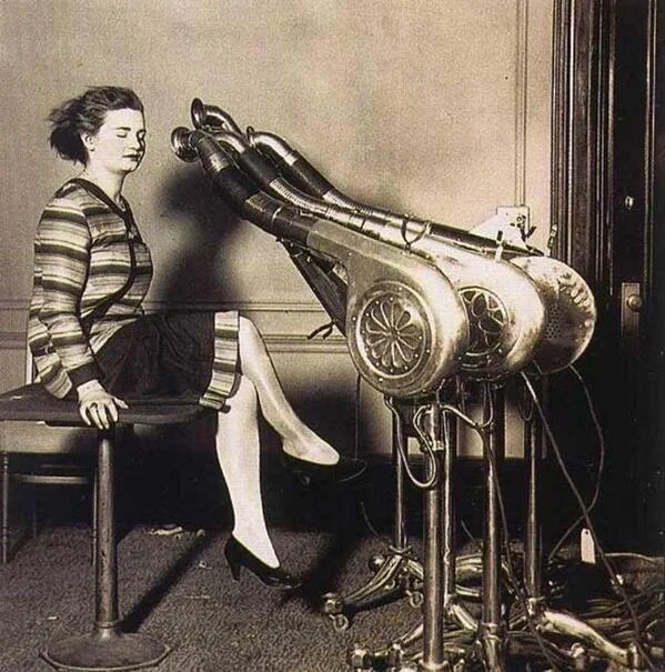 A woman sits with her eyes closed in front of a vintage hair drying machine with multiple metal hoses aimed at her head. She wears a striped dress and heels, and the room has a plain wall and wooden floor.