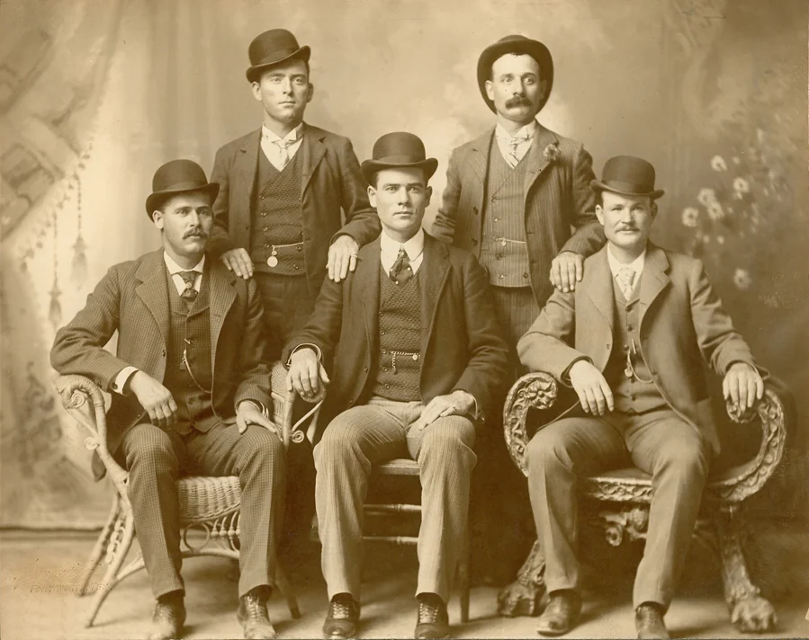 Five men in vintage suits and bowler hats pose for a sepia-toned portrait. Two men sit on chairs while three stand behind them. The background features a decorative curtain and flowers.