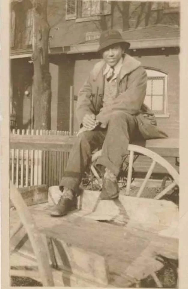 A person wearing a coat, hat, and boots sits on a wooden wagon outside a house with a picket fence. The scene appears to be historical, with sepia tones adding a vintage feel.