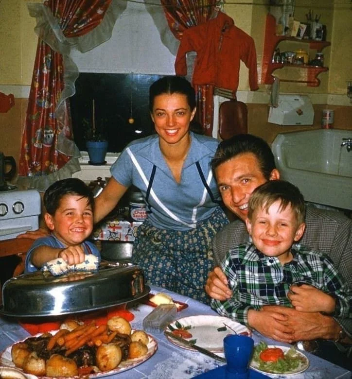 A smiling family of four gathers around a kitchen table filled with a meal. The mother stands behind, while the father hugs the two young boys, who are seated. The table is set with various dishes and a blue cup. The kitchen has vintage decor.