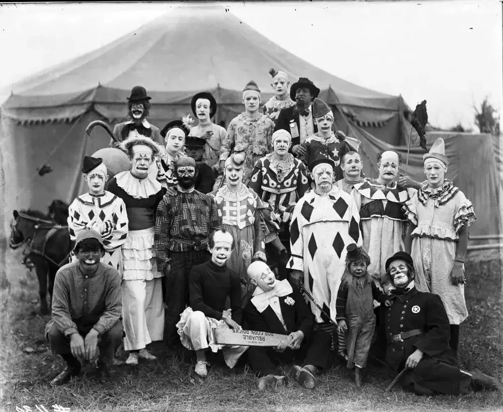 A black and white photo of a group of clowns in various costumes posing in front of a large circus tent. Some clowns wear polka dots and stripes, while others have hats or wigs. A small pony is seen on the left side.