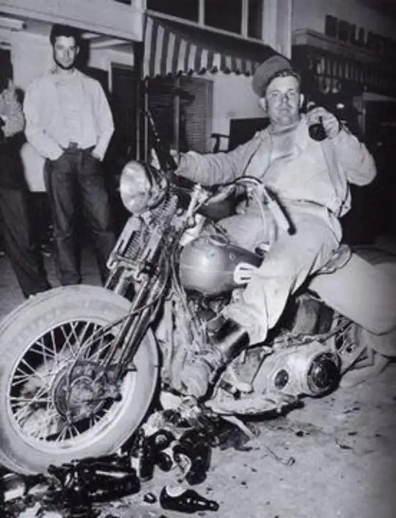 A man sits casually on a motorcycle in a street at night, holding a bottle. Broken glass is scattered on the ground beneath the bike. Another man stands nearby, leaning against a building with striped awnings. The photo is in black and white.