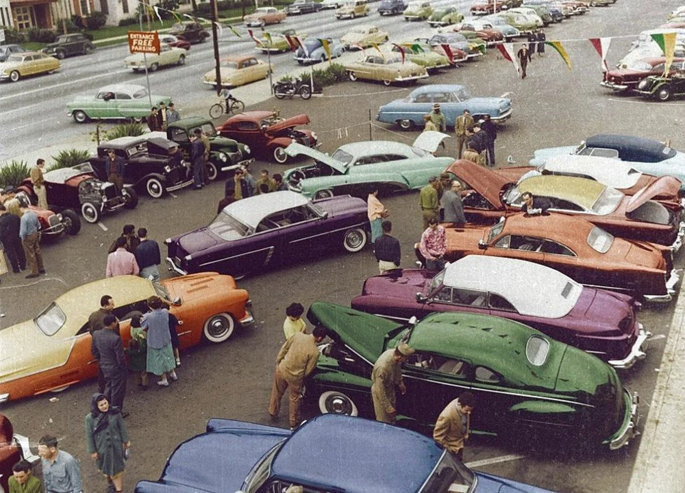 A bustling 1950s car dealership lot with colorful vintage cars parked and numerous people browsing. The scene is lively, with a street in the background where more cars are passing by. Banners and a "Free Parking" sign are visible.