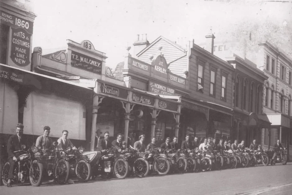 A historic black and white photo shows a line of motorcycles parked on a street with riders standing beside them. Behind them are old shops and buildings, including a butcher and a clothing store. The scene evokes an early 20th-century feel.