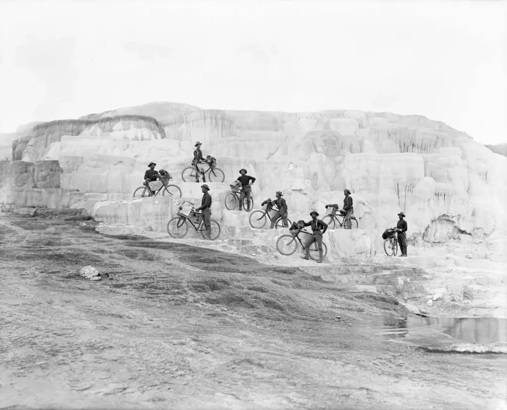 A group of people stands with bicycles on the terraced formations of Mammoth Hot Springs in Yellowstone National Park. The white, layered rock formations contrast with the dark clothing of the cyclists. The scene is a historic black and white photograph.