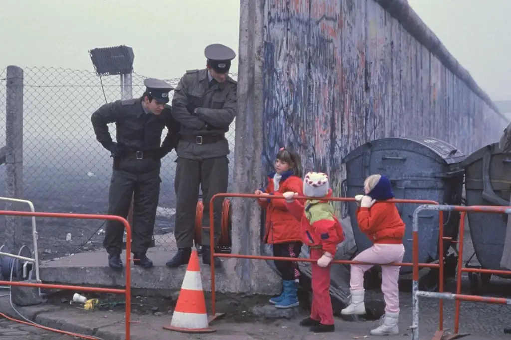 Two uniformed guards stand facing three children near a graffiti-covered wall. The children are dressed in bright winter clothes and are by a red barrier. The backdrop includes a chain-link fence and a foggy sky.