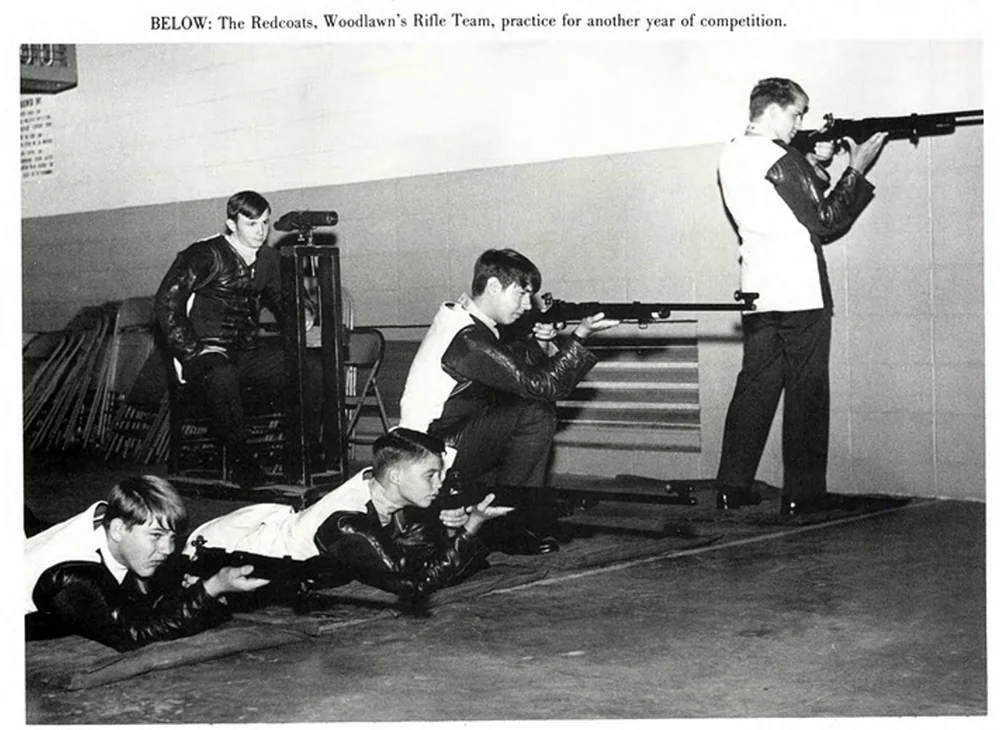 A group of five young people, part of Woodlawn's Rifle Team, practice shooting. Four are lying on mats aiming rifles, while one stands to aim. They wear matching jackets in a gym-like setting with stacked equipment in the background.