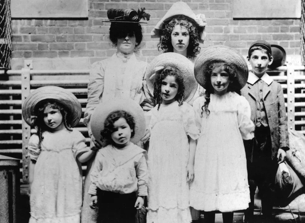 A vintage black-and-white photo of a group. Two adults in hats stand behind five children, four girls in white dresses and one boy in a suit. They are posed outside in front of a brick wall and a wooden fence.