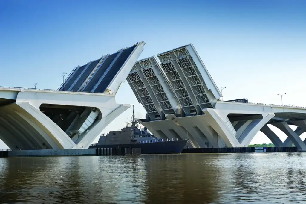 A drawbridge with two sections raised vertically, allowing a navy ship to pass through beneath. The bridge spans a calm body of water under a clear blue sky.