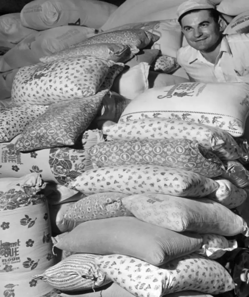 A smiling man wearing a cap stands behind a large stack of patterned flour sacks. The sacks are piled high, each with distinct designs and labels. The scene appears to be in a storage area filled with numerous similar-filled sacks.