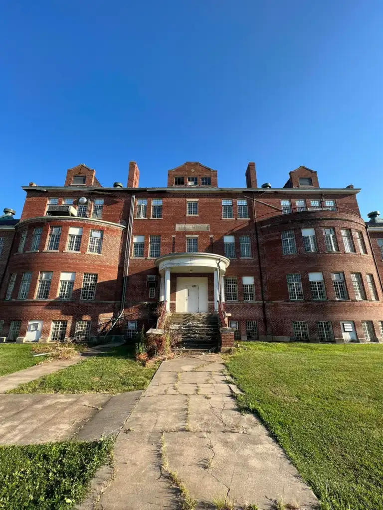 A large, historic red brick building with multiple windows and chimneys under a clear blue sky. The structure has a prominent central entrance with stone steps, surrounded by a grassy lawn and a paved walkway leading up to it.