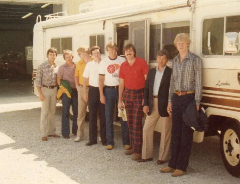 A group of eight men are standing in front of a white recreational vehicle. They are casually dressed in various shirts and pants, some in jeans and plaid. The background shows a garage area with an open door.