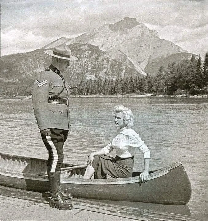 A person in a Royal Canadian Mounted Police uniform stands on a dock, looking at a woman seated in a canoe. They are by a lake with a mountain in the background. The scene is in black and white.