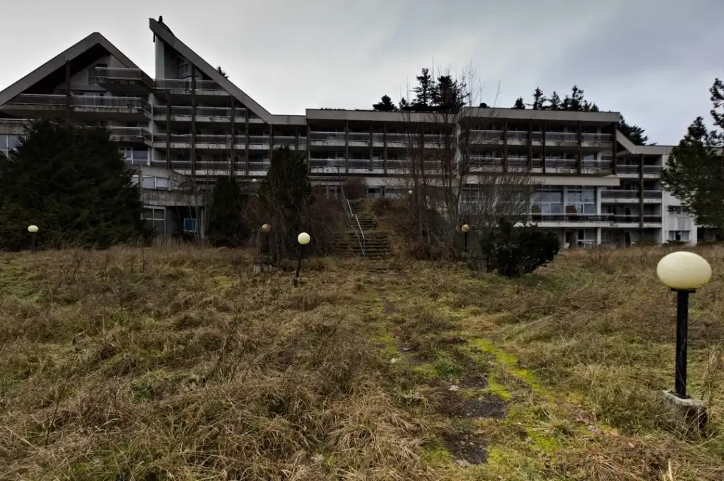 Overgrown grass and a path lead to an abandoned, multi-story building. The structure is composed of geometric shapes with balconies. A few globe streetlights line the path, and trees surround the area under a cloudy sky.