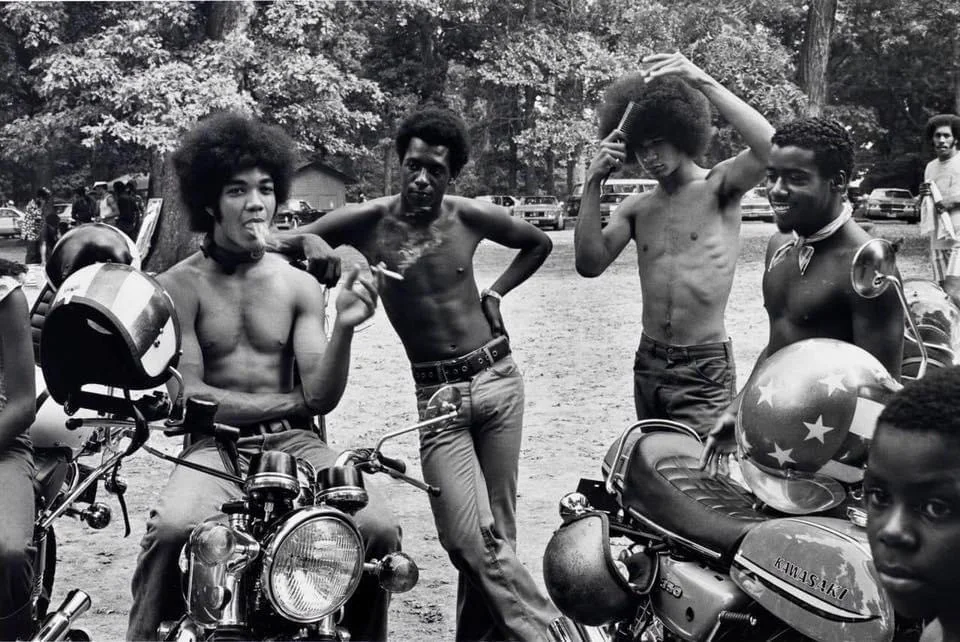 A group of five young men pose with motorcycles in a shaded outdoor area. Some are shirtless, and two are seated on bikes. Trees and parked cars are visible in the background. One motorcycle helmet has an American flag design.