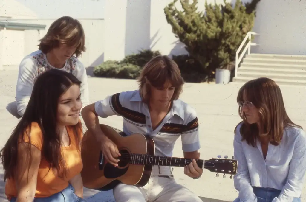 A group of four young people sitting outdoors. One person is playing an acoustic guitar, while the others listen and engage. The scene is sunny with trees and a building in the background. The mood is casual and relaxed.