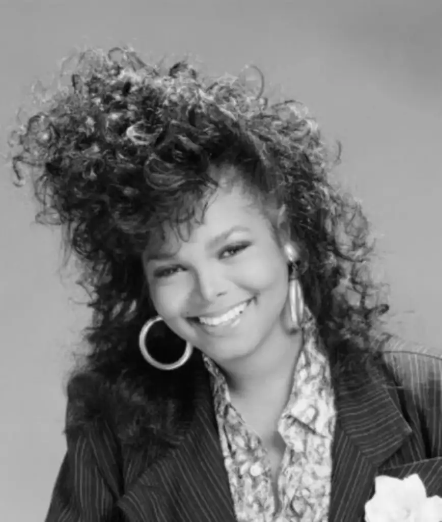 Black and white portrait of a smiling woman with voluminous curly hair and hoop earrings. She is wearing a patterned blouse under a pinstripe blazer.