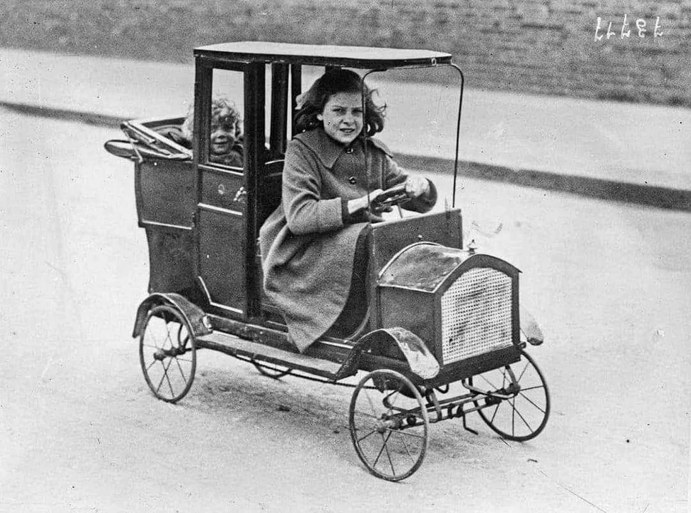 A black-and-white photo of a young girl driving a small, vintage, toy car on a street. The car has a classic design with thin wheels and an open-top. A young boy is seated behind her, visible through the back window.