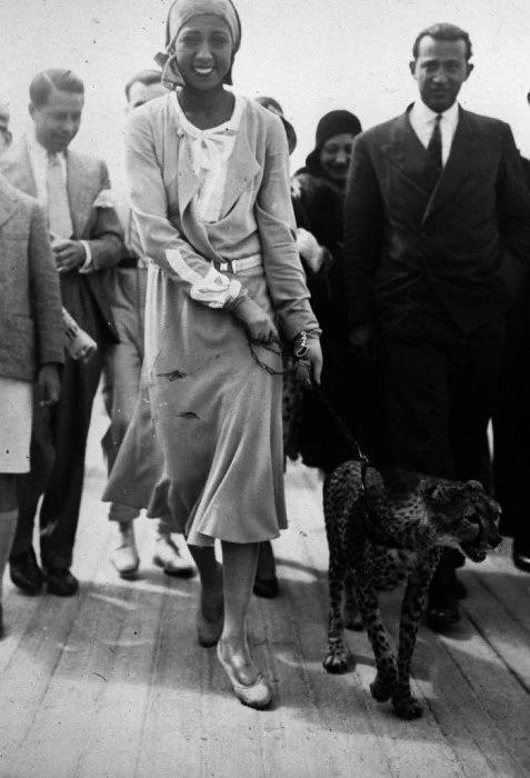 A woman in a 1920s-style dress and headscarf walks a small leopard on a leash, surrounded by a group of men and women on a wooden floor. The scene appears joyful and lively.