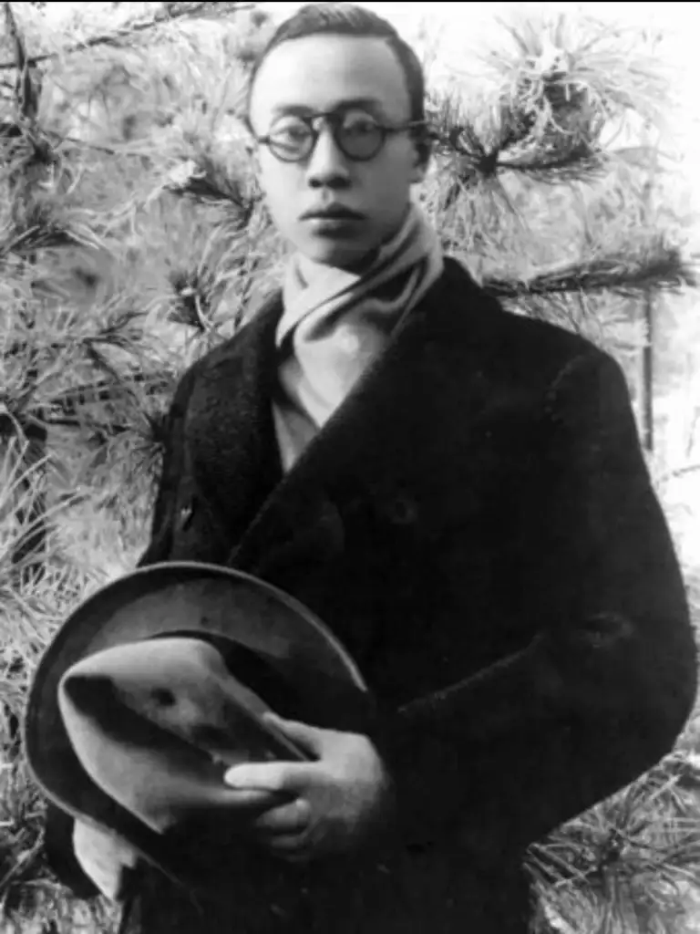 A person wearing glasses and a dark overcoat stands outdoors beside a tree with sparse branches. They hold a hat and scarf, and the background is filled with pine needles. The photo has a vintage black-and-white aesthetic.