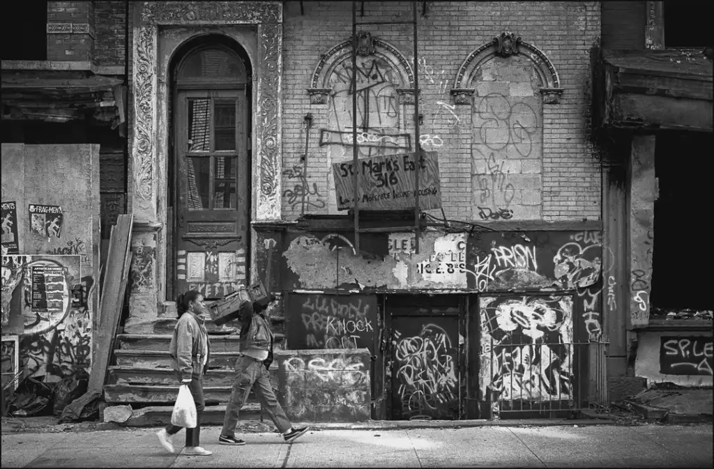 Two people walk past an old, graffiti-covered building with boarded windows and crumbling brickwork. One holds a plastic bag, and the other gestures with an arm. The atmosphere is gritty and urban.