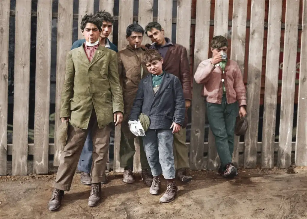 Six boys stand in front of a wooden fence. They are dressed in worn, mismatched clothing. One boy holds a cigarette, while others have hands in pockets or hold caps. The setting appears to be outdoors on a dirt surface.