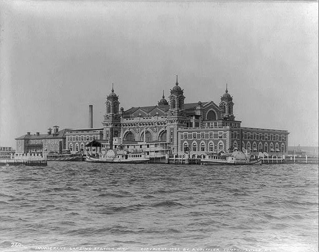 Black and white photo of the large, historic Ellis Island Immigration Station building surrounded by water. The structure has a symmetrical design with multiple arches and towers. Several boats are docked at the pier in the foreground.