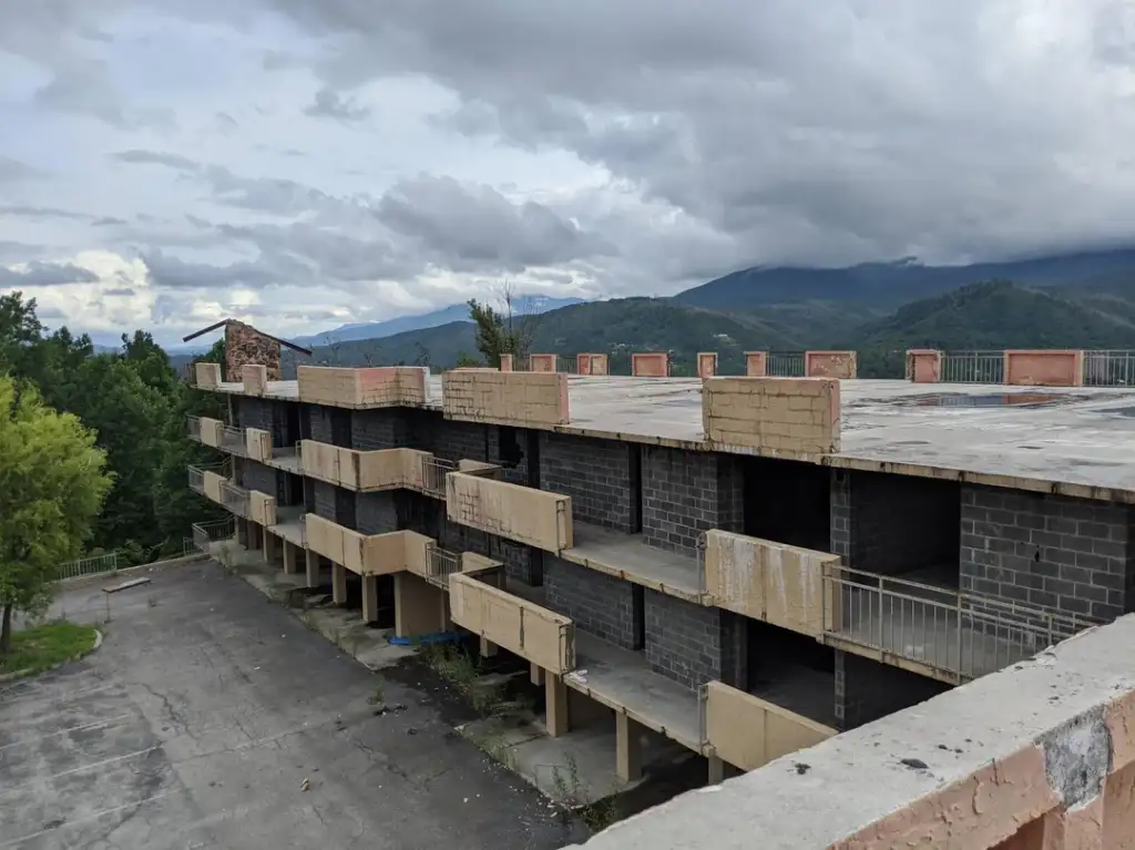 Abandoned multi-story building with unfinished balconies, surrounded by trees and mountainous terrain under a cloudy sky. The structure has exposed concrete and brickwork on its exterior.