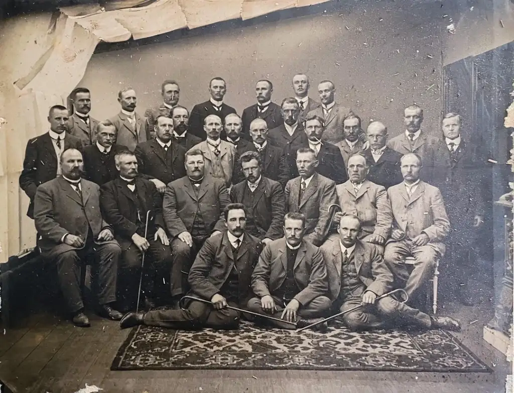 A vintage black and white photo of a group of men in formal attire, posing in three rows; some seated and others standing. The backdrop is plain, and they are on a patterned rug. The image has a sepia tone and shows signs of aging.