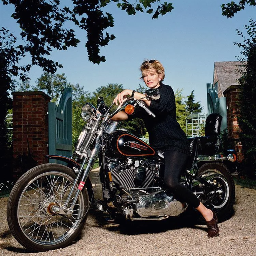 A person with short hair sits on a Harley-Davidson motorcycle, wearing a black outfit. They are parked on a gravel path, with a brick wall and open gate in the background, and trees surrounding them. The sky is clear and blue.