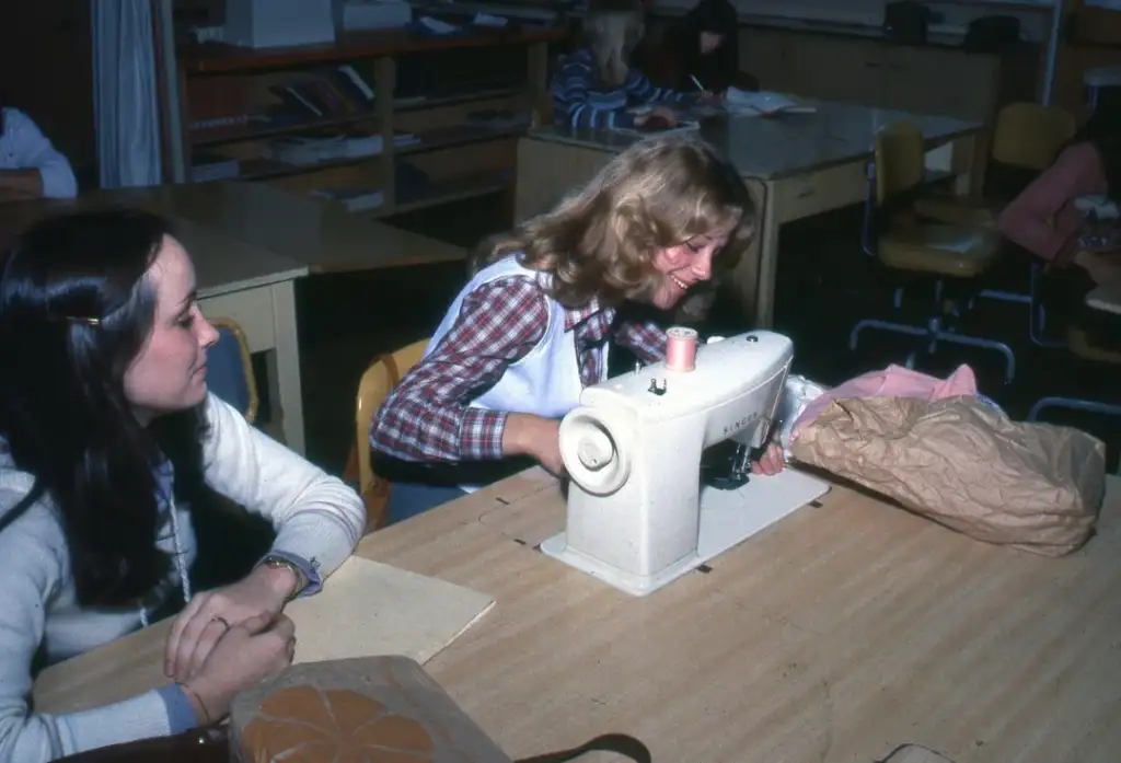 A blonde woman in a plaid shirt uses a sewing machine at a wooden table, while a brunette woman in white observes. Bookshelves and more people are visible in the background.