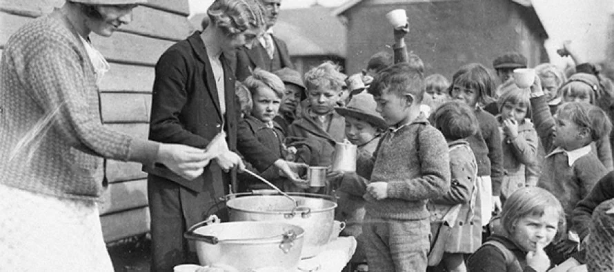 A historical black-and-white photo of children and adults gathered around large pots during a feeding program. A woman serves porridge to a young boy among a group of children holding cups. The setting appears outdoors near a building.